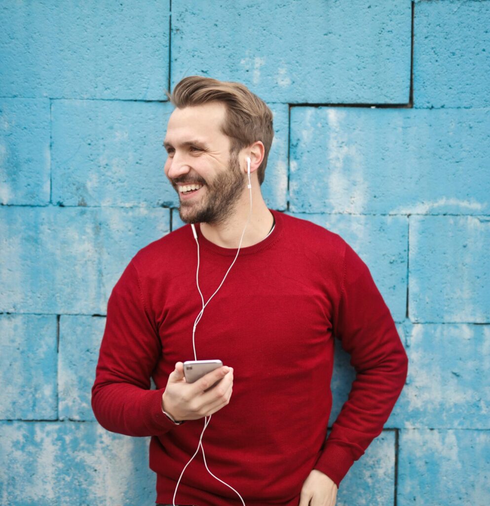 A joyful man in a red sweater smiles while listening to music on his smartphone against a blue brick wall.